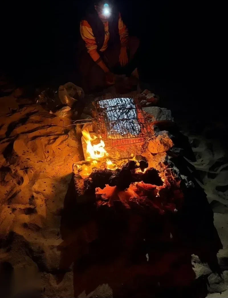 A guide grilling meat on a wire rack over glowing campfire coals at night.
