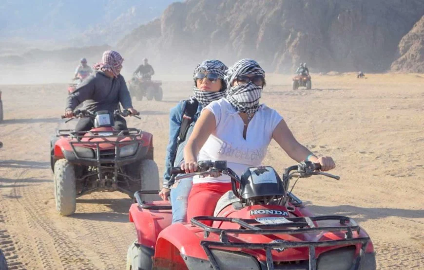 Two women wearing shemagh scarves riding a quad bike together through the desert, with other riders following behind.