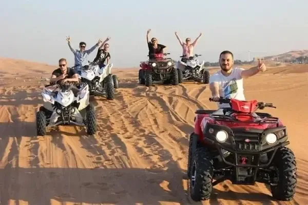 Group of six riders on quad bikes cheering with arms raised on a sandy desert trail.