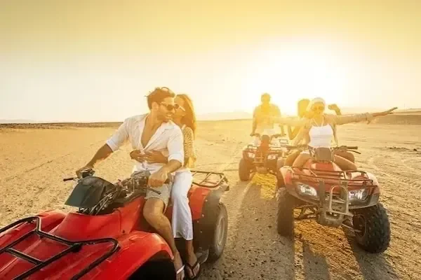 A couple sits together on a red quad bike in warm desert light, with another pair on a quad bike behind them.