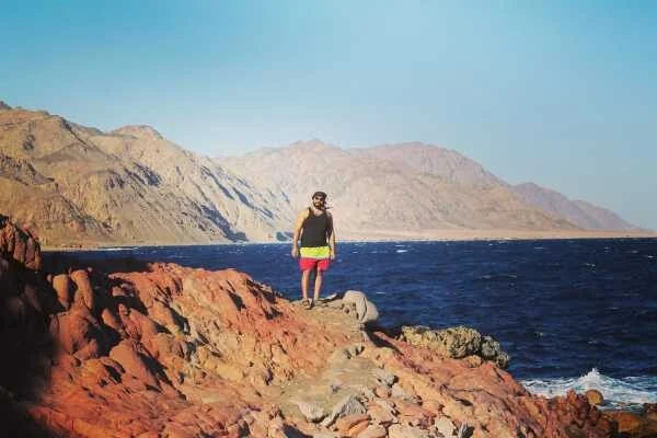 A man in swim shorts stands on red rocky coastline overlooking the blue sea, with mountains lining the shore.