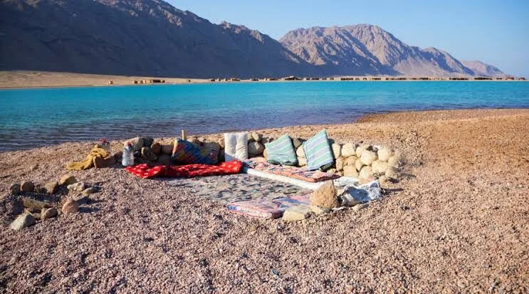 Colorful cushions and mats are arranged on a pebble beach beside a turquoise lagoon, with mountains and beach huts in the distance.