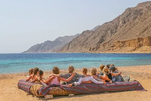 A group of people lie together on a striped mat on the beach, looking out at the turquoise lagoon and mountains.