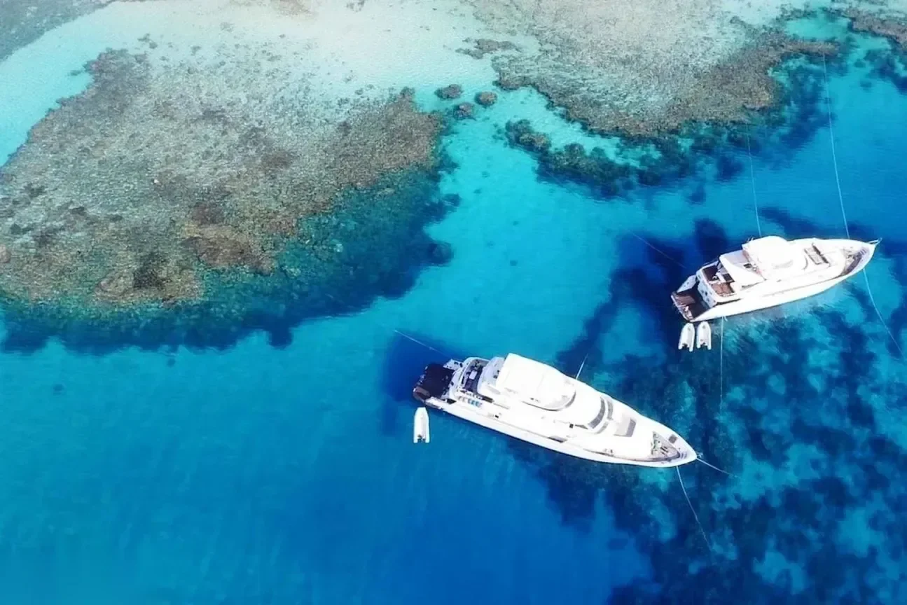 Aerial view of two motor yachts anchored in clear turquoise water beside patches of coral reef.