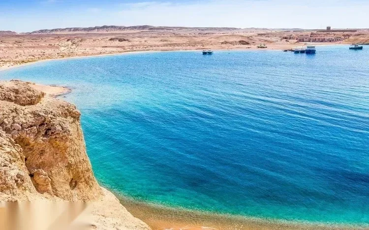 Elevated view of a curved turquoise bay with a rocky headland, sandy beach, and small boats moored in the distance.
