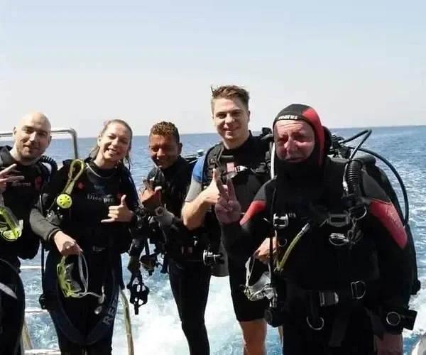 A group of five divers pose for a photo on the back of a boat, giving thumbs-up and hand signals.