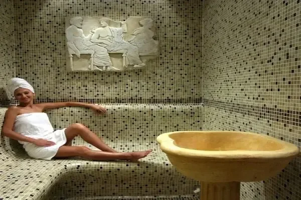 Woman wrapped in a white towel with a towel turban, reclining on a heated marble bench beside a carved stone basin under a classical relief carving