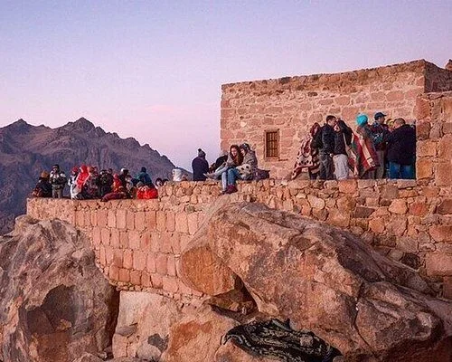 Hikers gathered around a small stone summit building on Mount Sinai at dusk with mountain ranges in the distance