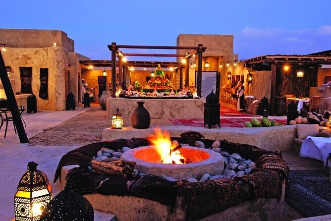 Desert Bedouin-style camp at dusk with a stone fire pit, lanterns, decorated tables and lounge seating around mudbrick buildings