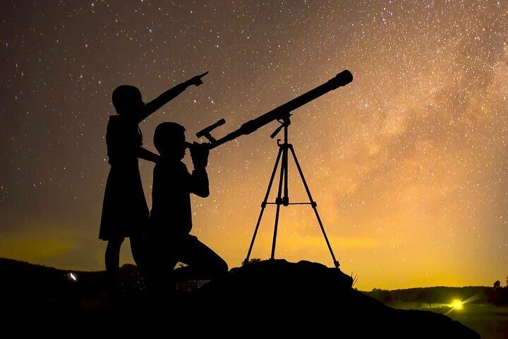 Silhouette of two people using a telescope on a tripod against a glowing star-filled sky with the Milky Way visible