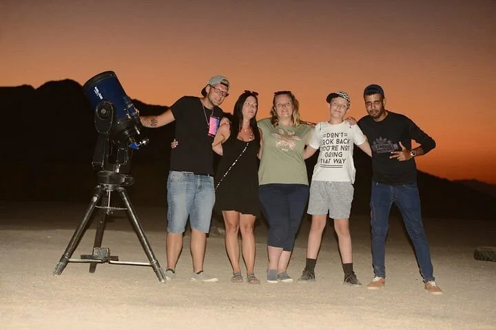 Group of five people posing beside a large telescope on a tripod in the desert at sunset with mountains behind