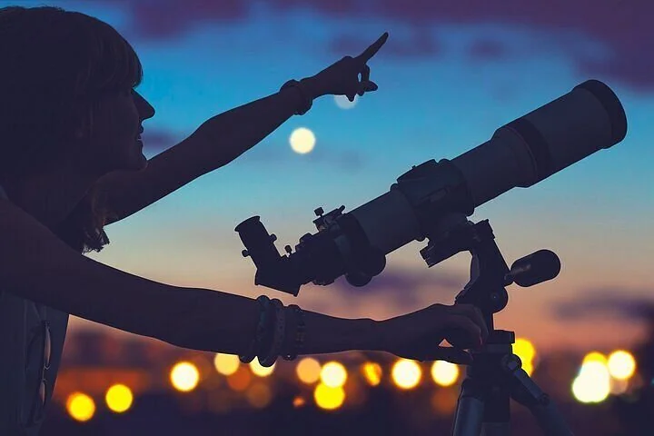 Silhouette of a woman pointing skyward beside a telescope with the moon and stars visible against a fading sunset sky