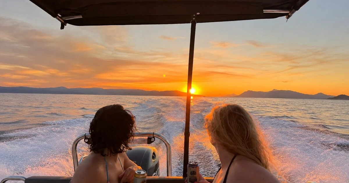 Two women relaxing on the back of a moving speedboat holding drinks as the sun sets over the hills