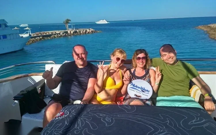 Four friends sitting on a yacht's bow smiling for a photo, harbor with anchored boats and a rock breakwater in the background, one holding a branded 'Marsa Alam Tours' towel