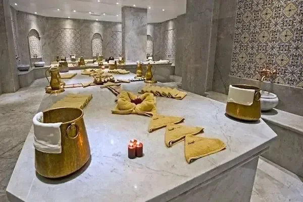 Marble hammam table set with folded gold towels arranged in a decorative pattern, brass buckets, and lit candles