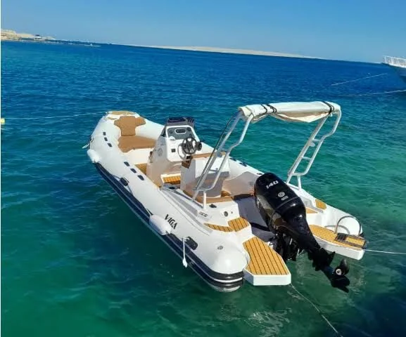 White rigid inflatable speedboat with tan cushioned seating and an outboard motor, anchored in clear turquoise water near a sandy shore