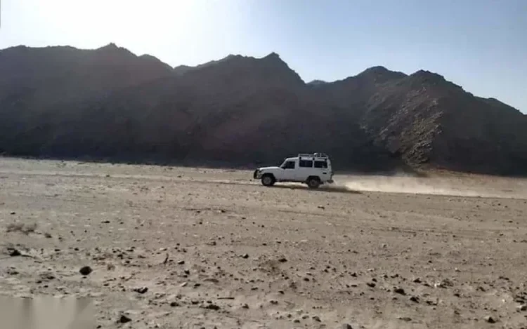 White 4x4 jeep speeding across an open desert plain kicking up a trail of dust with mountains in the distance
