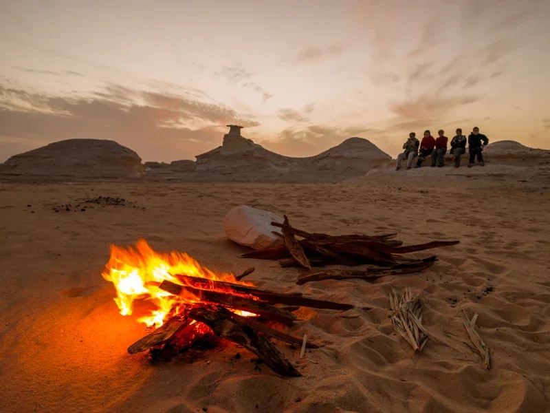 Campfire burning on the sand at sunset with a group of tourists sitting on white rock formations in the background
