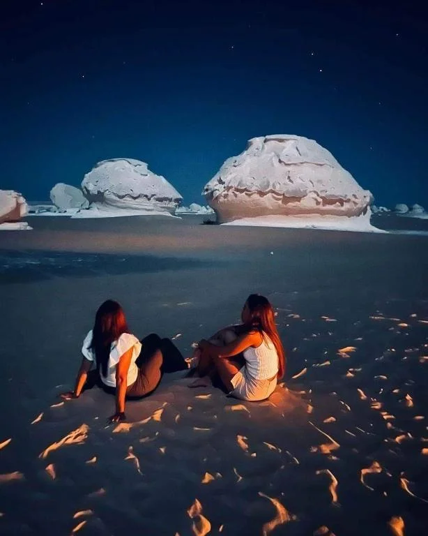 Two women sitting on the sand at night beside glowing white rock formations under a starry sky