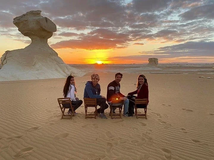 Four tourists sitting on camp chairs watching the sunset beside a mushroom-shaped rock formation in the desert