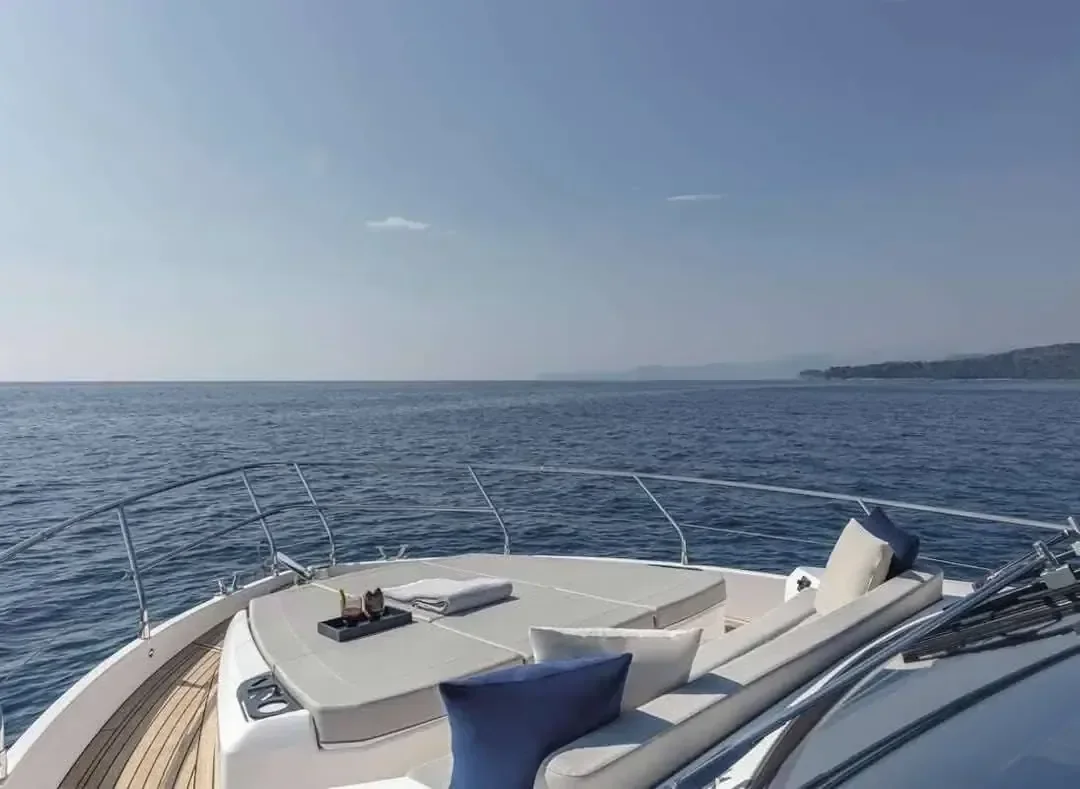 Empty bow deck of a white luxury yacht with cream cushioned loungers and navy blue pillows, calm sea and hazy coastline in the background