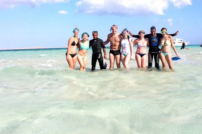 Group of eight tourists and two dive guides standing in shallow turquoise water wearing snorkel masks and fins, a boat visible behind them