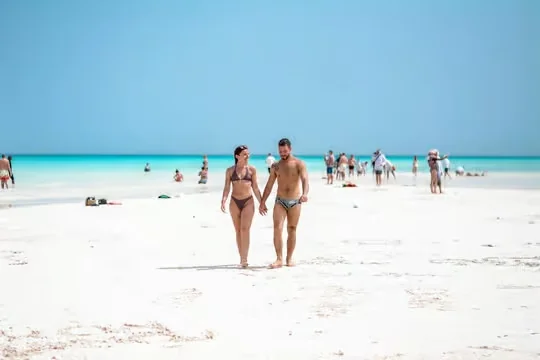 Couple walking hand in hand along a white sand beach beside turquoise water with other beachgoers in the background