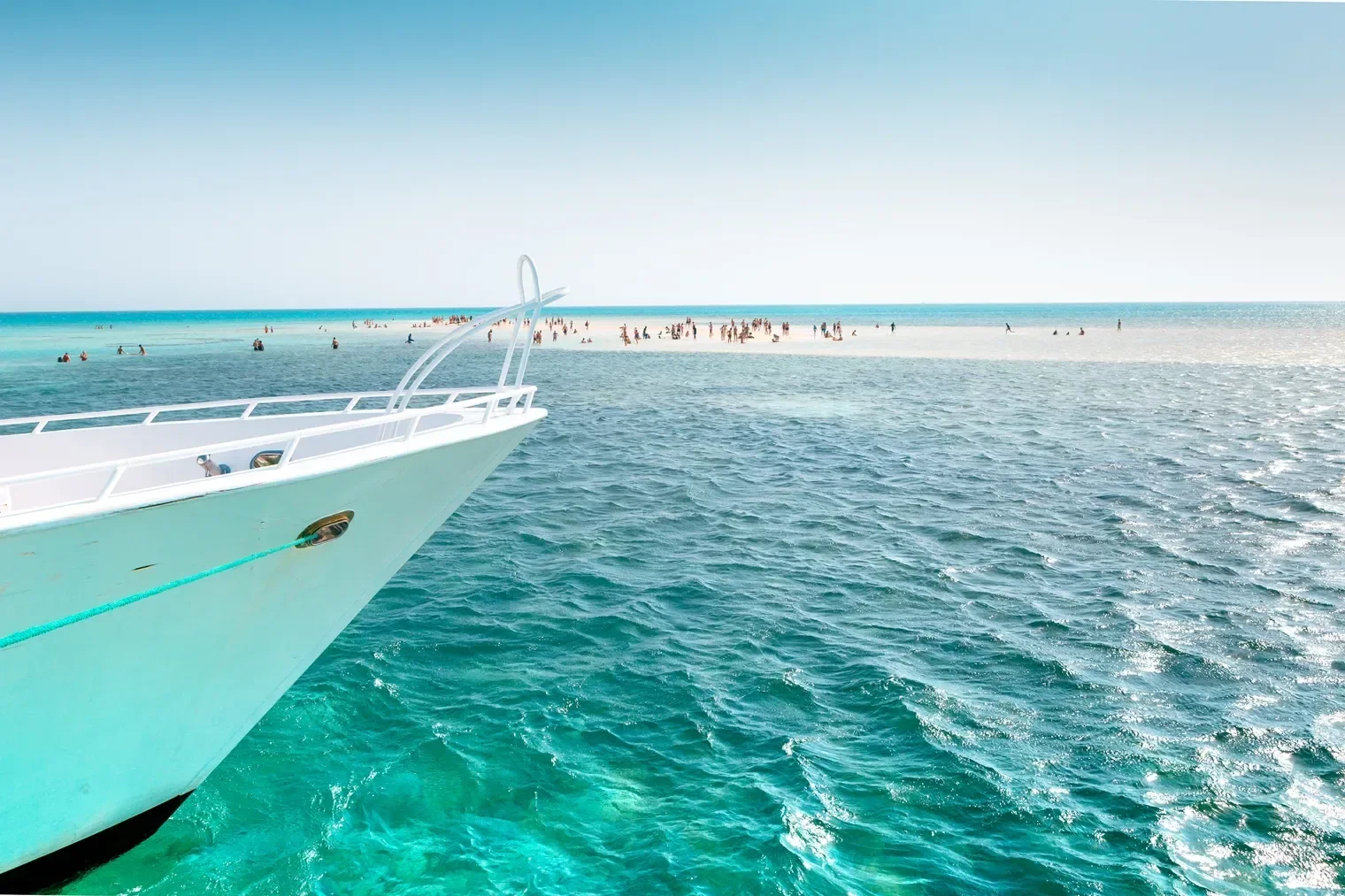 View from a yacht's bow with a turquoise rope leading toward a crowded white sandbar island where beachgoers wade in shallow water